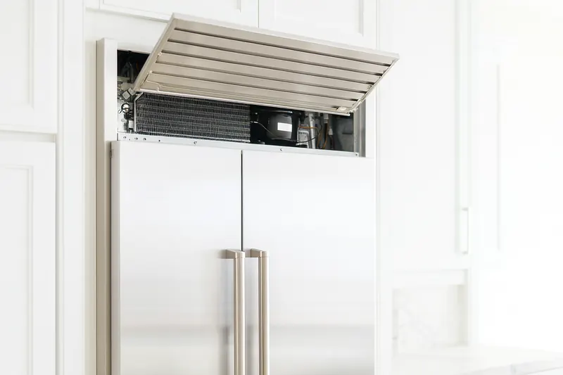 Top machine compartment of a built-in refrigerator with the grille removed, showing the condenser coil and fan above the doors