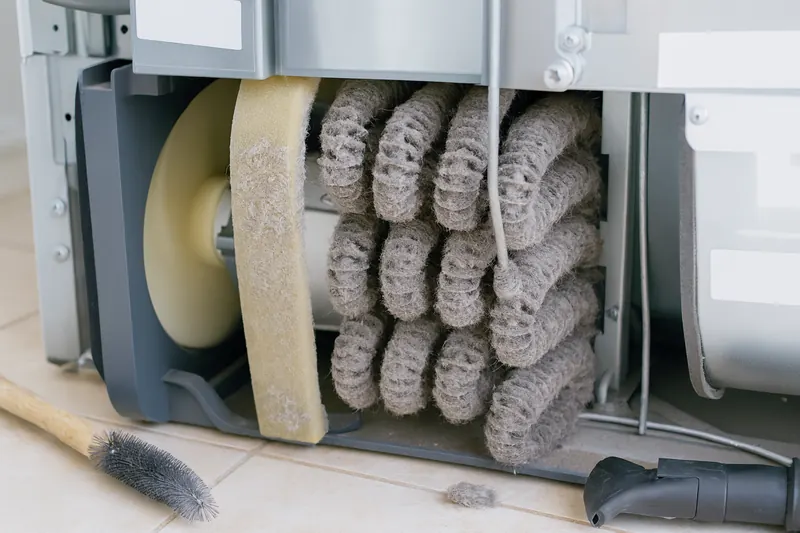 Refrigerator condenser coil heavily coated with gray dust and lint behind a removed bottom grille